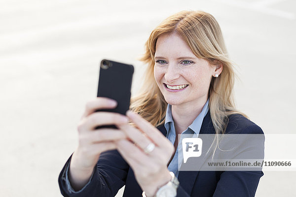 Porträt einer lächelnden  blonden Geschäftsfrau  die sich mit dem Smartphone selbstständig macht.