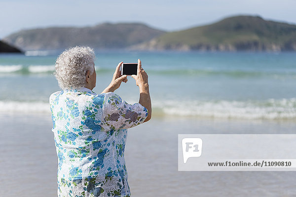 Back view of senior woman on the beach taking picture with smartphone