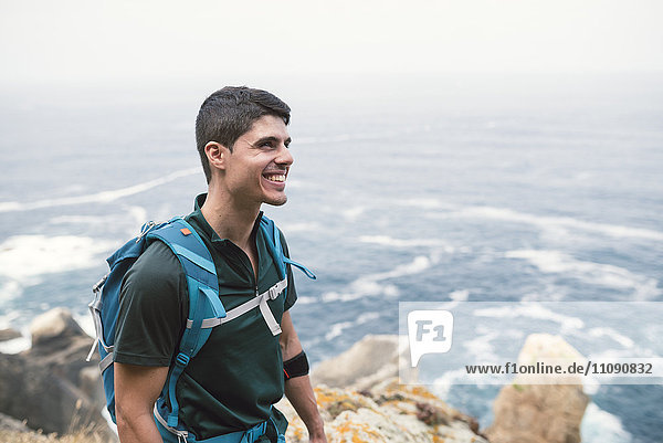 Lächelnder Mann mit Rucksack vor dem Meer an der Küste