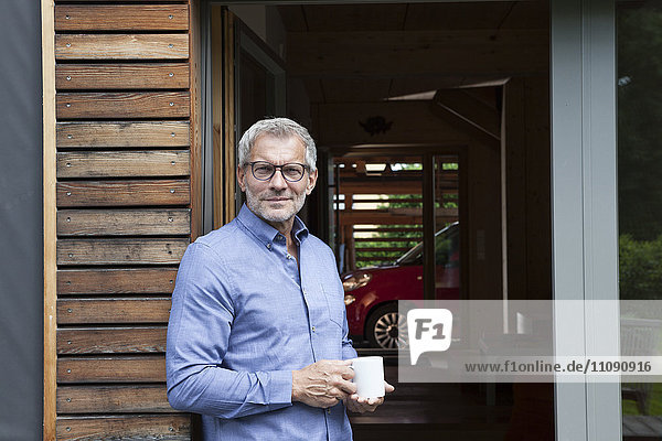 Mature man holding cup leaning against terrace door