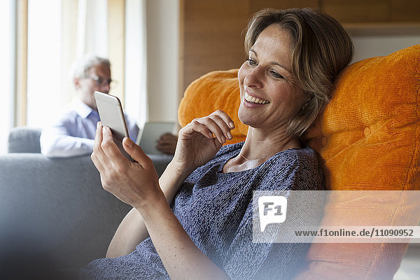 Lächelnde Frau zu Hause beim Blick auf das Handy mit Ehemann im Hintergrund