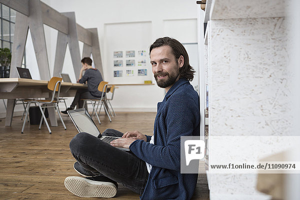 Smiling man sitting on floor in office working on laptop