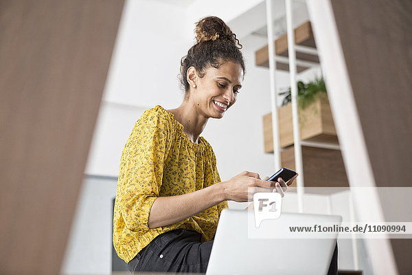 Smiling woman sitting on office desk using cell phone