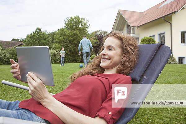 Lächelnde Frau im Garten mit Tablette und Familie im Hintergrund