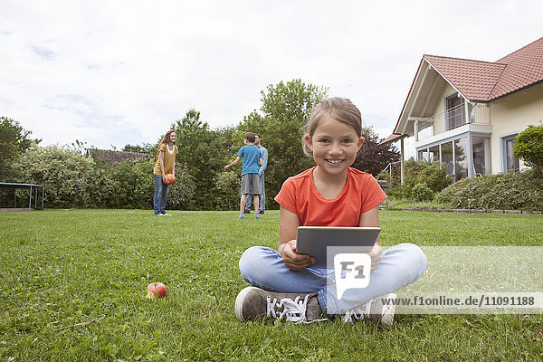 Lächelndes Mädchen im Garten sitzend mit Tablette und Familie im Hintergrund