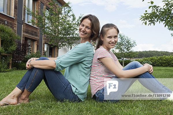 Smiling mother and daughter sitting back to back in garden
