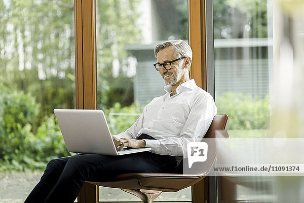 Lächelnder Mann sitzt auf Stuhl in seinem Wohnzimmer mit Laptop