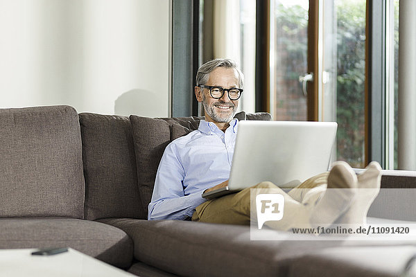 Lächelnder Mann sitzt auf der Couch in seinem Wohnzimmer mit Laptop