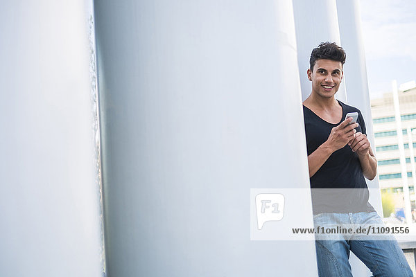 Lächelnder junger Mann mit an der Säule lehnendem Smartphone