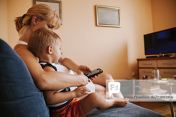 Pregnant mother and her little son sitting on the couch with remote control