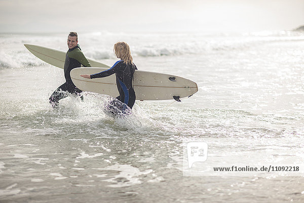 Paar mit Surfbrettern  die zusammen durch die Wellen laufen.