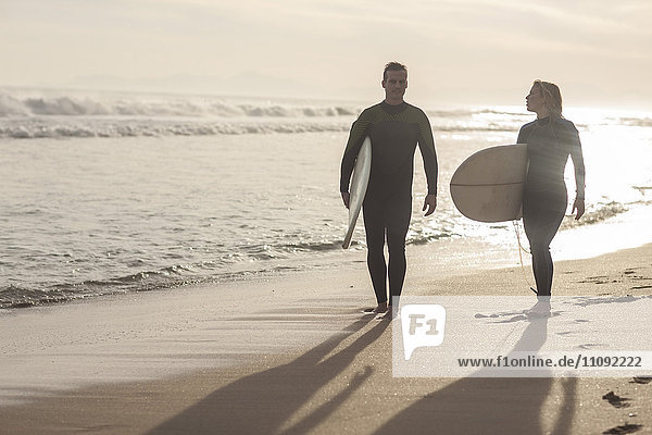 Paar mit Surfbrettern  die bei Sonnenuntergang am Strand spazieren gehen.