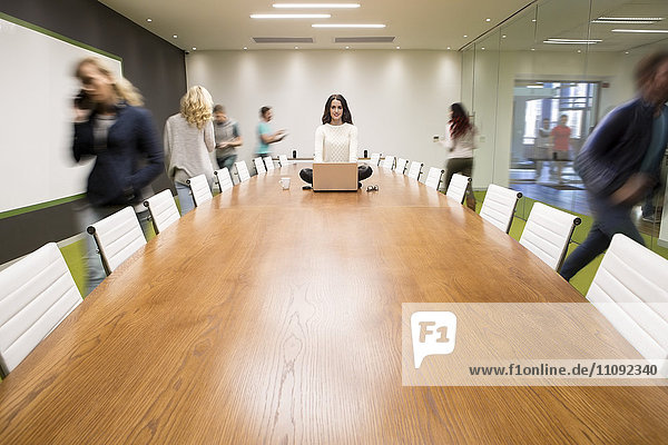 Businesswoman with laptop sitting on conference table with people running around
