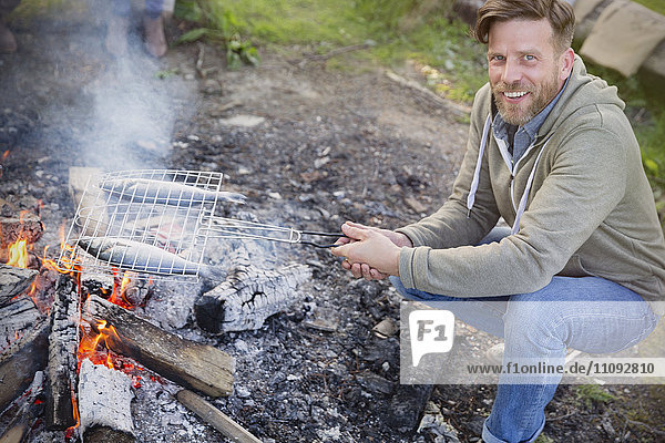 Portrait lächelnder Mann beim Kochen von Fisch im Grillkorb am Lagerfeuer