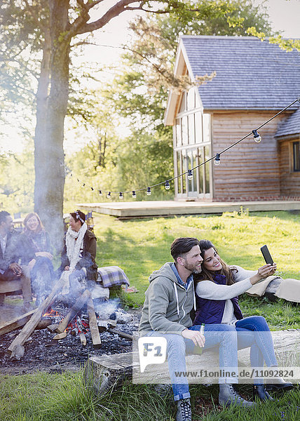 Couple taking selfie with camera phone at campfire outside cabin