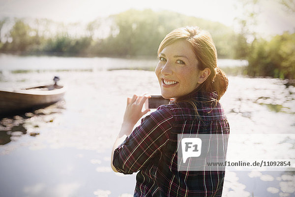 Portrait lächelnde Frau mit roten Haaren fotografiert sonnigen See mit Fotohandy