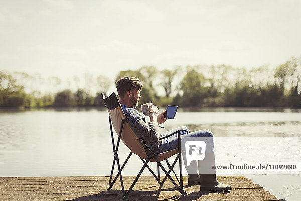 Mann trinkt Kaffee und benutzt digitales Tablett am sonnigen Seeuferdock