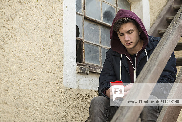 Junger Mann schreibt Textnachrichten auf seinem Smartphone und sitzt auf einer Holztreppe