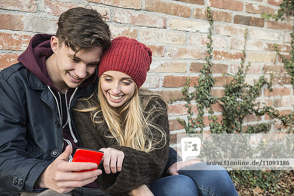 Junges Paar lächelt und benutzt ein Smartphone an einer Mauer