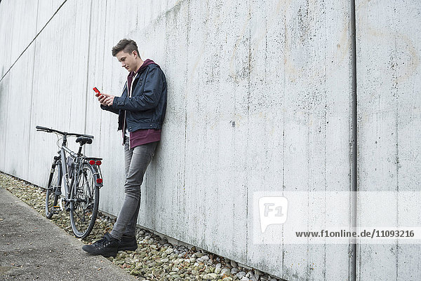 Junger Mann schreibt eine SMS auf seinem Smartphone und lehnt sich an eine Betonwand