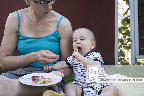 Kleiner Junge isst Kuchen vom Teller seiner Mutter auf dem Rasen  München  Bayern  Deutschland