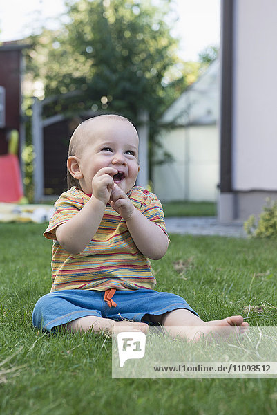Kleiner süßer Junge sitzt auf Gras und lacht auf einer Wiese  München  Bayern  Deutschland