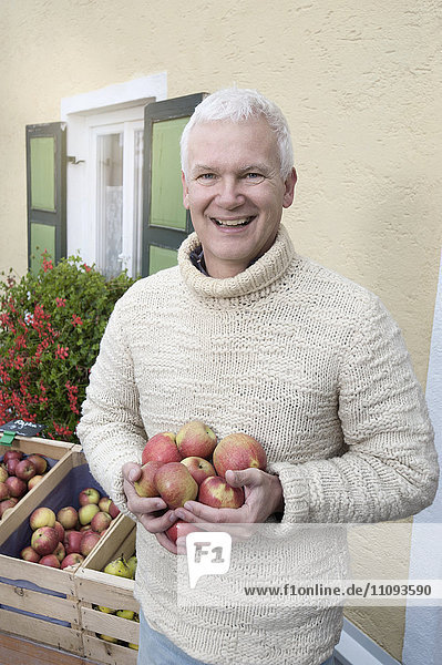Porträt eines reifen Mannes  der vor einem Bioladen Äpfel in den Händen hält und lächelt