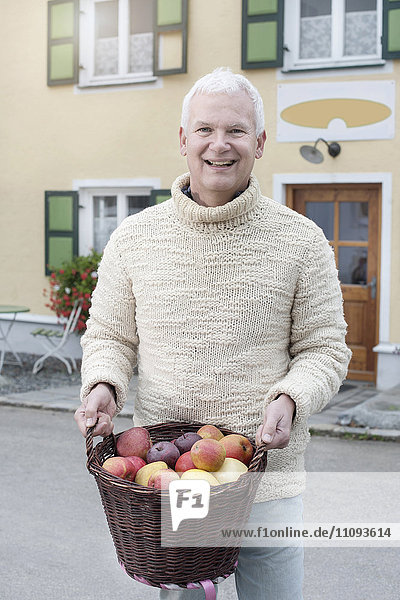 Porträt eines reifen Mannes  der lächelnd einen Korb voller Äpfel in den Händen hält  vor einem Bioladen