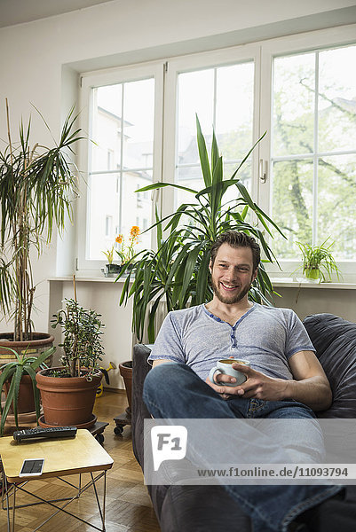 Mid adult man drinking coffee in living room and smiling  Munich  Bavaria  Germany