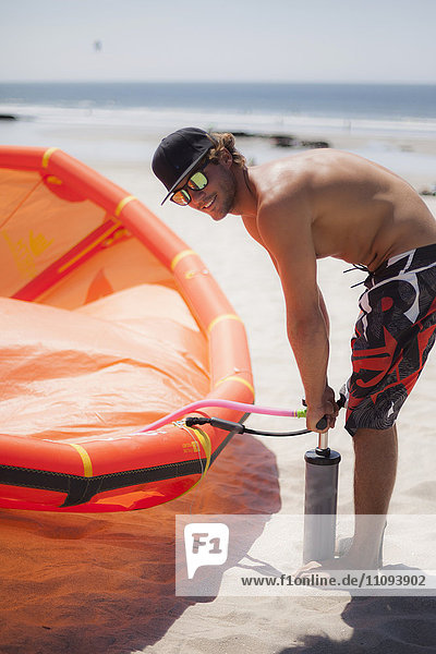 Junger Mann pumpt Luft in ein Kiteboard am Strand