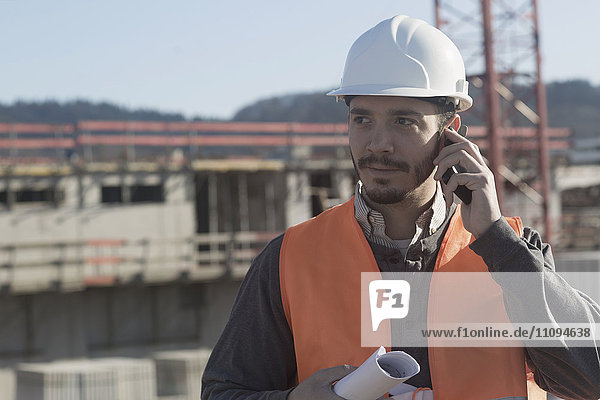 Junger männlicher Ingenieur,  der einen Bauplan hält und auf einer Baustelle mit einem Mobiltelefon spricht