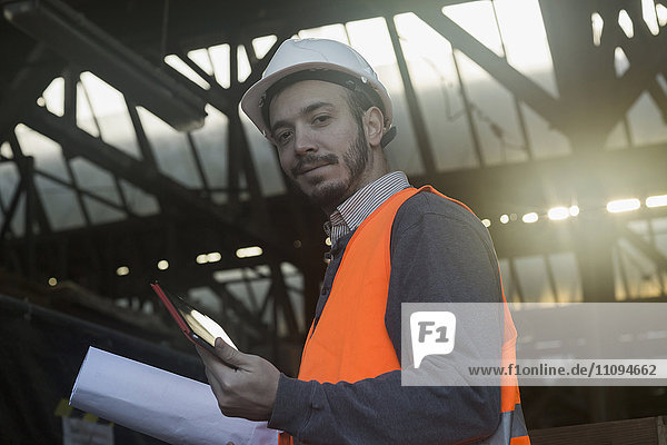 Porträt eines jungen männlichen Ingenieurs  der einen Bauplan hält und ein digitales Tablet auf einer Baustelle benutzt