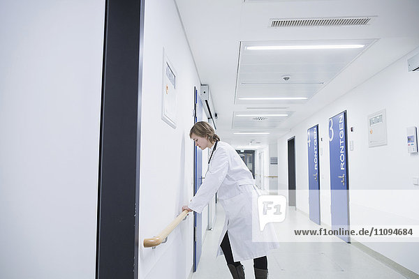 Seitenprofil einer überforderten jungen Ärztin,  die sich in einem Krankenhauskorridor an ein Geländer lehnt