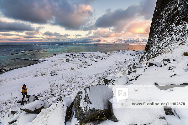 Fotograf auf dem Schnee bewundert das Fischerdorf unter einem bunten Himmel Eggum  Vestvagoy (Vest-Vagoy) Insel  Lofoten Inseln  Arktis  Norwegen  Skandinavien  Europa