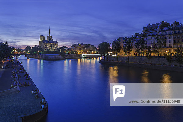 Gruppen von Parisern und Touristen säumen die Ufer der Seine mit der in der Abenddämmerung beleuchteten Notre Dame auf der Ile de la Cite  Paris  Frankreich  Europa