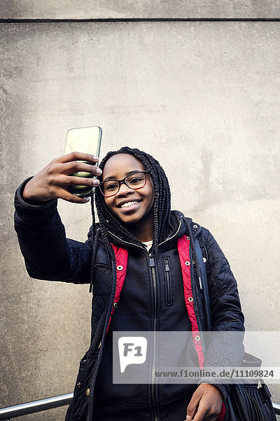 Low angle view of smiling teenager taking selfie through smart phone against wall