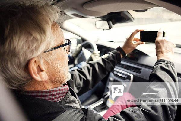 Senior Mann fotografiert mit Smartphone durch die Windschutzscheibe des Autos