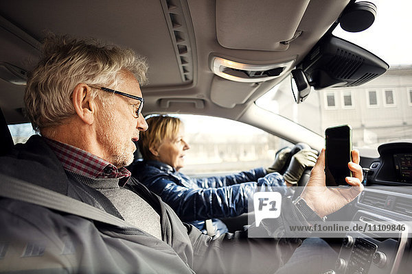Mann hält Smartphone  während er von einer älteren Frau beim Autofahren in der Stadt sitzt.