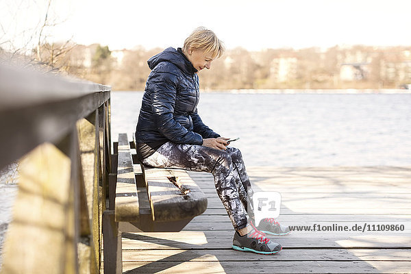 Senior woman in sportswear using phone while sitting on bench by lake