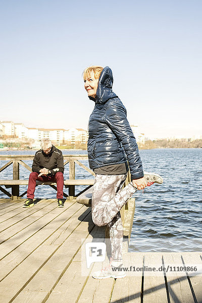 Ältere Frau streckt das Bein  während der Mann auf der Bank am Pier gegen den klaren Himmel sitzt.