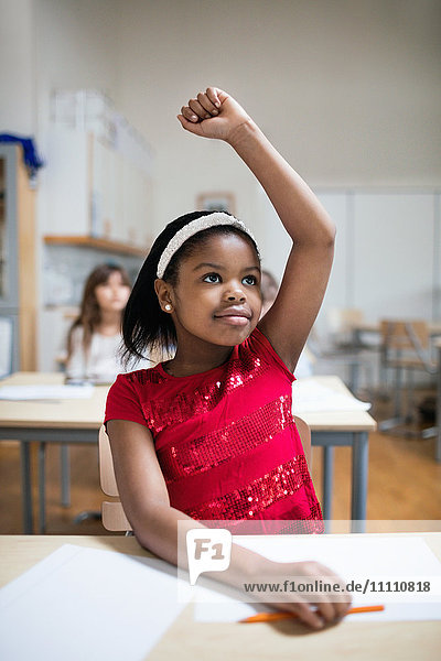 Konzentriertes Mädchen mit erhobener Hand am Schreibtisch im Klassenzimmer sitzend
