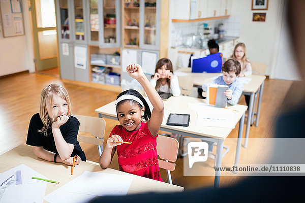 Lächelnder Schüler mit erhobener Hand im Klassenzimmer sitzend