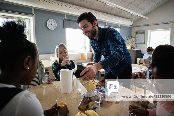 Teacher serving juice to students sitting at table in classroom