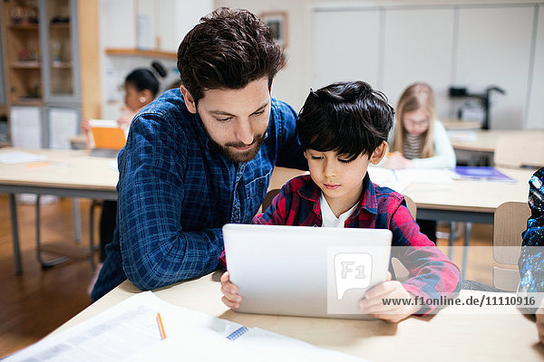 Lehrer  der den Schüler bei der Benutzung des digitalen Tabletts im Klassenzimmer unterstützt.