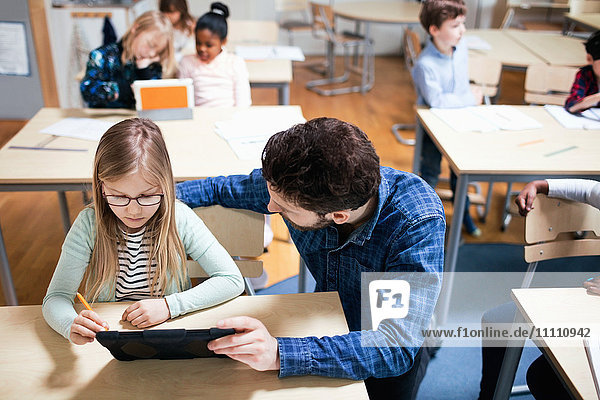 Hochwinkelansicht des Lehrers  der die Schülerin bei der Verwendung des digitalen Tabletts im Klassenzimmer unterstützt.