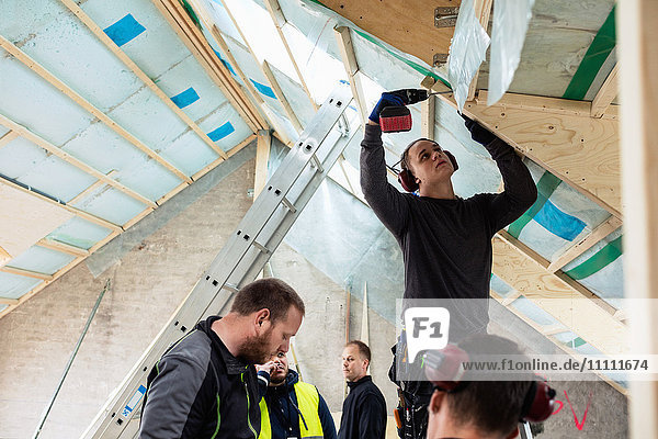Bauarbeiter beim Bohren des Dachbalkens  während die Mitarbeiter auf der Baustelle an der Leiter stehen