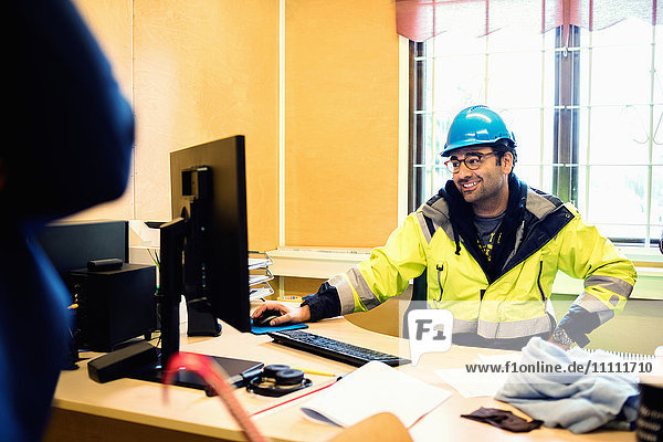 Lächelnder Bauarbeiter in Schutzkleidung im Gespräch mit dem Mann am Schreibtisch