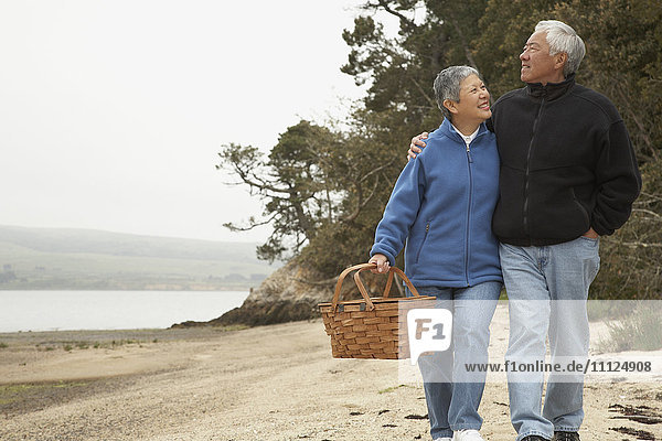 Älteres asiatisches Paar geht mit Picknickkorb am Strand spazieren