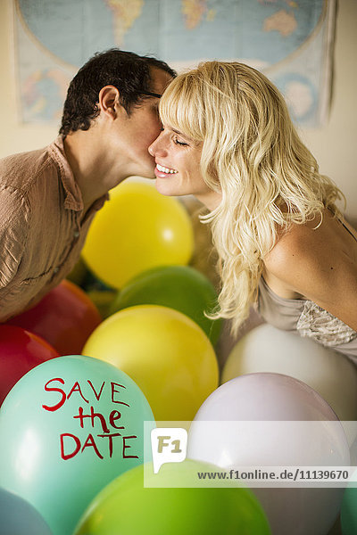 Kaukasisches Paar küsst sich über einem Luftballon mit dem Text 'save the date''.