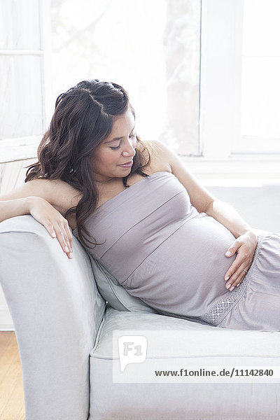 Pregnant Hispanic woman holding belly on sofa